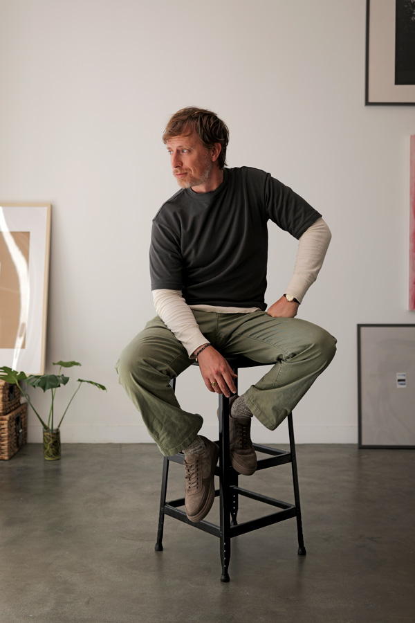 Man seated on a stool wearing a dark t-shirt over a cream long sleeve thermal with olive pants and brown suede sneakers