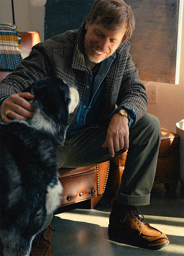 Man in the Station Jacket seated and petting a black and white dog, wearing brown moc toe boots